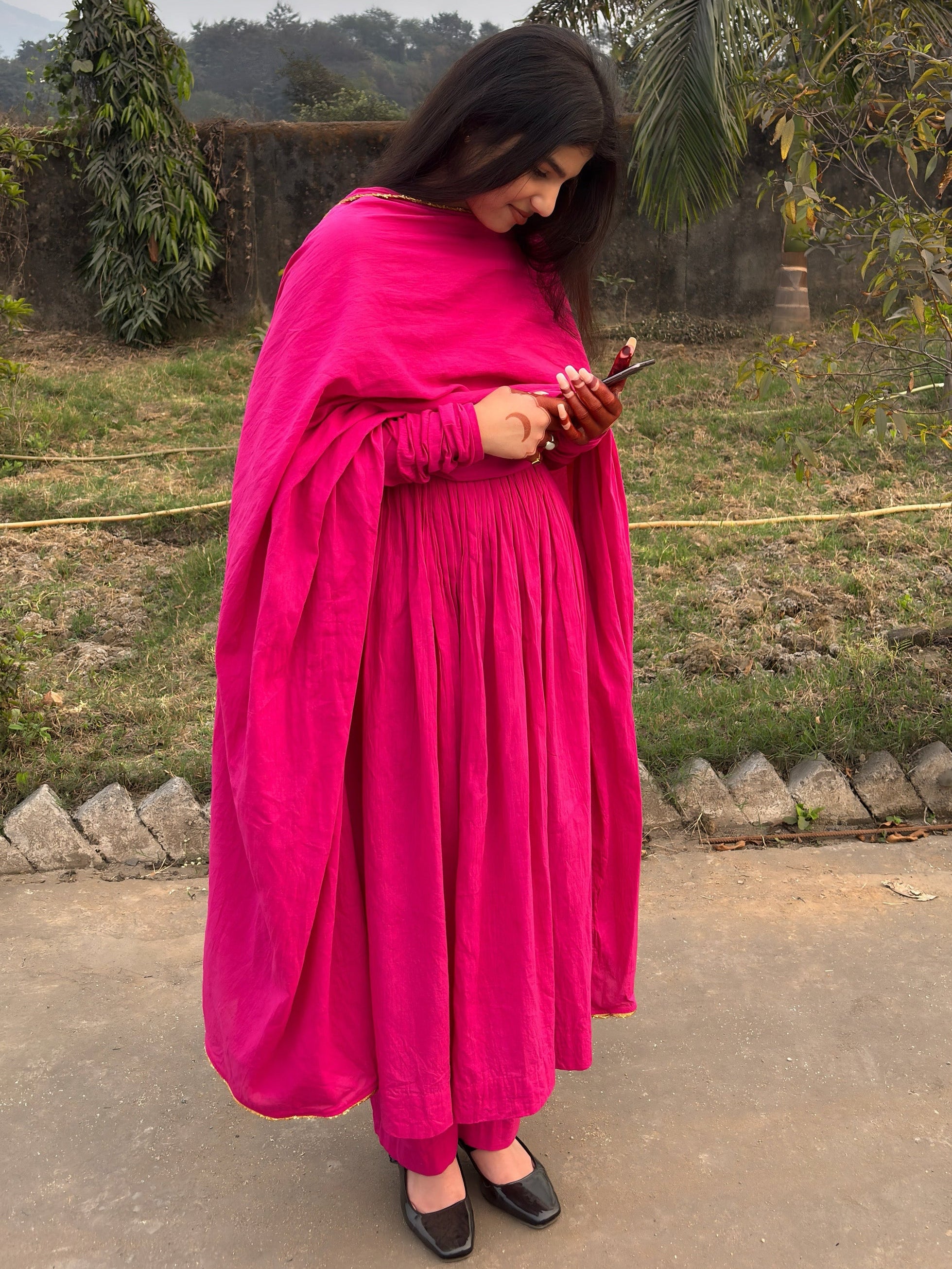 Woman in a bright pink traditional outfit standing outdoors with trees and a fence in the background.